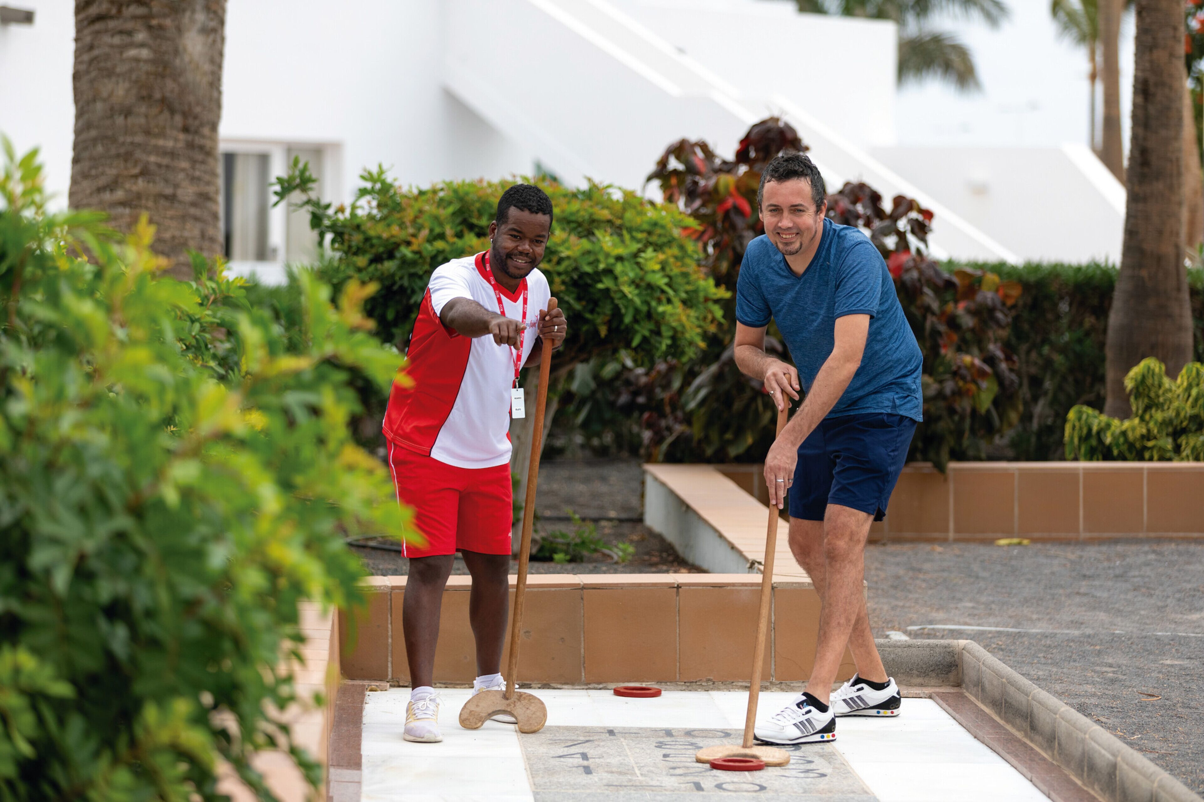 Two people playing shuffleboard on an outdoor court with palm trees and a white building in the background.
