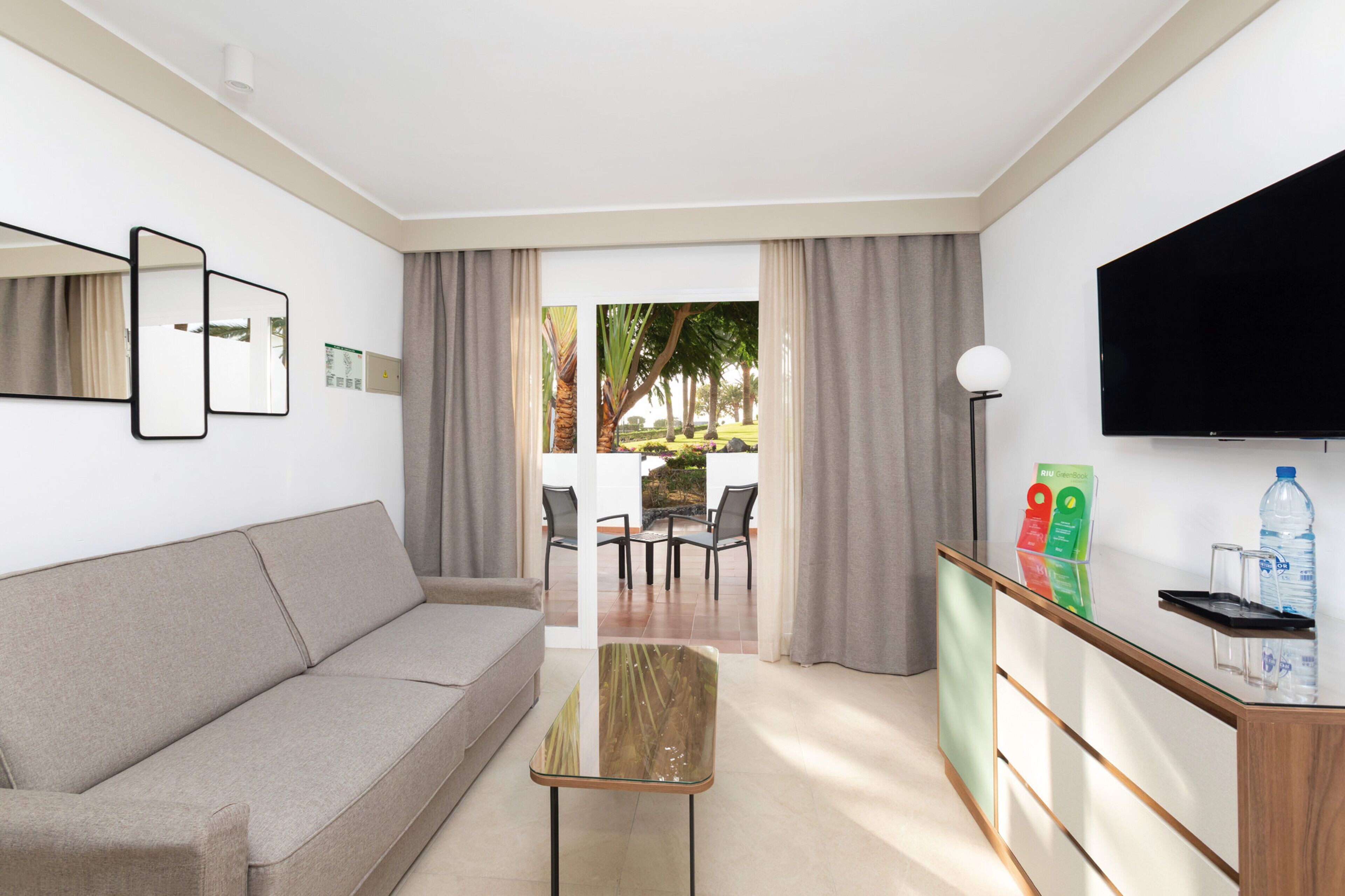 Una sala de estar moderna con un sofá, una mesa de centro de vidrio, un televisor de pantalla plana y vista a un patio exterior con sillas y una mesa.