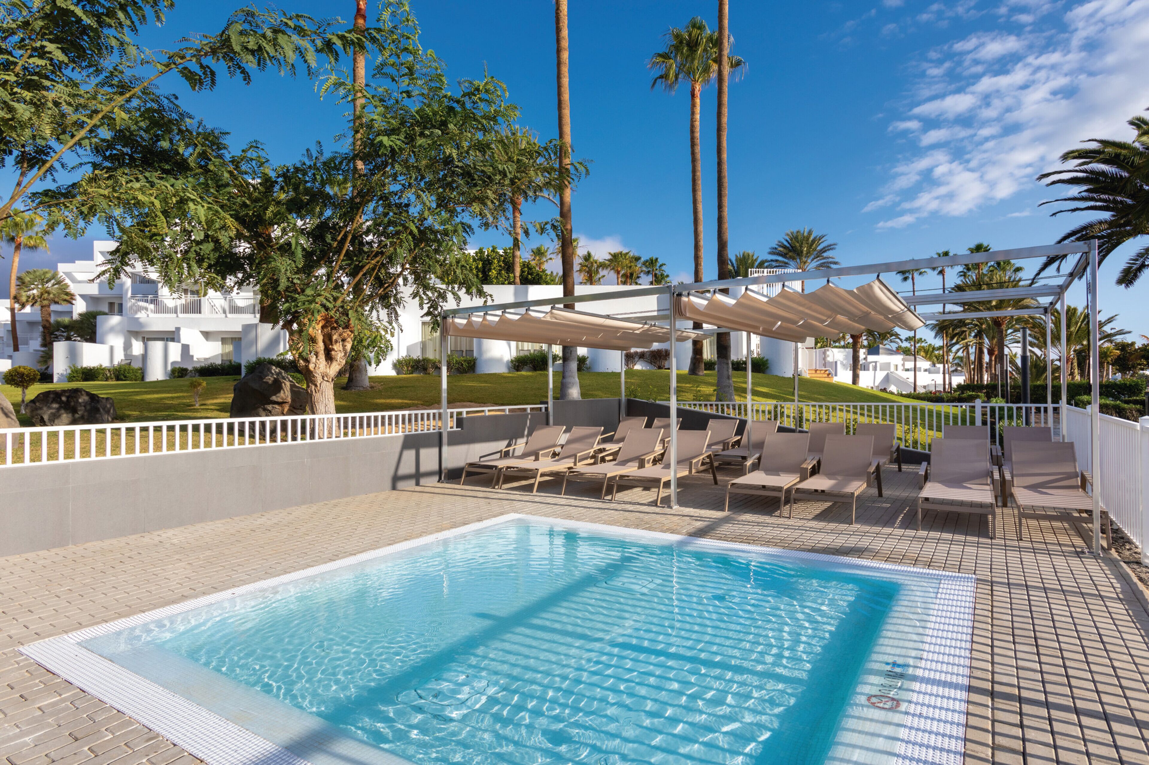 A swimming pool with lounge chairs and a covered area, surrounded by palm trees and a white building.