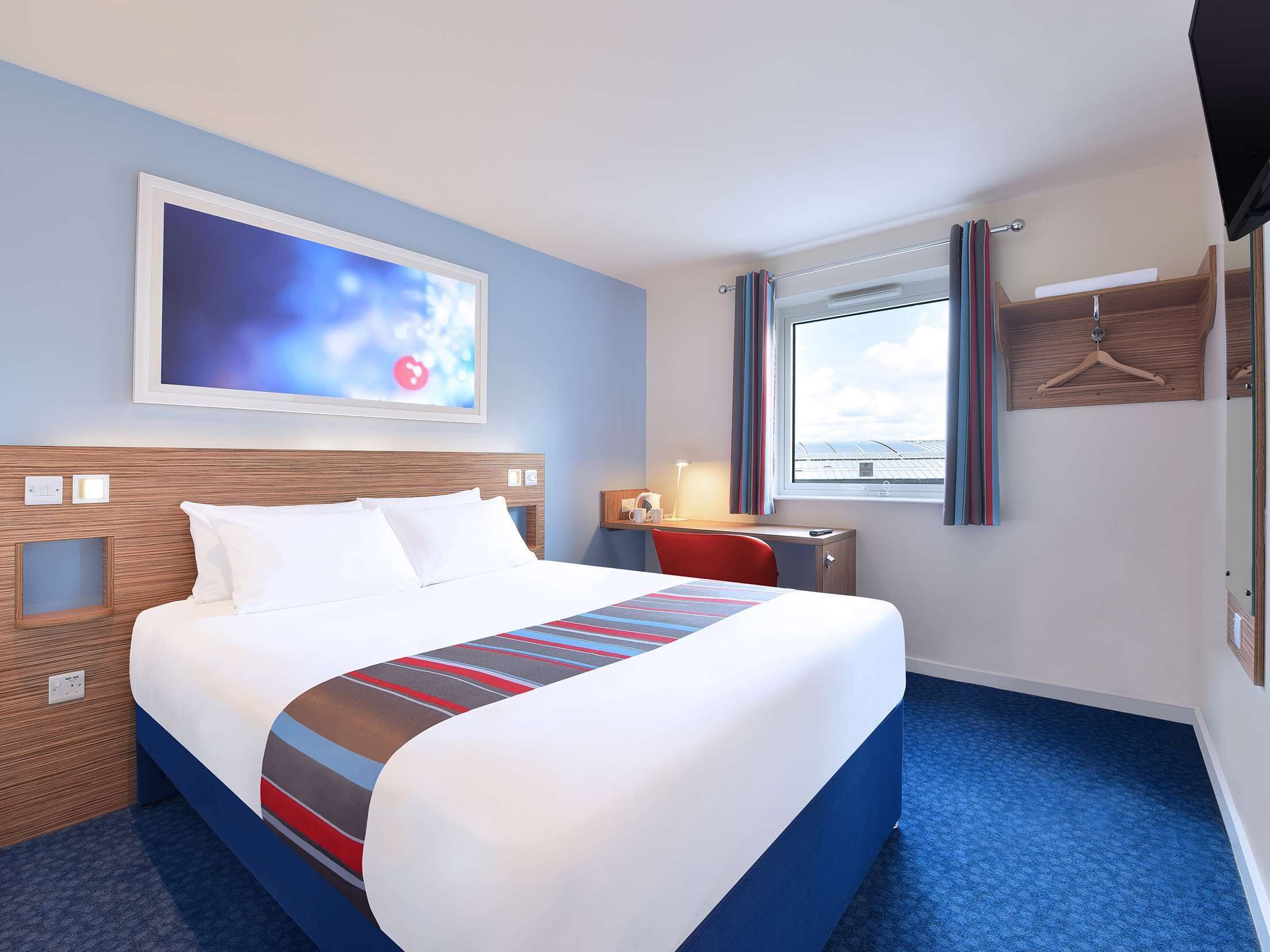 Quarto de hotel com cama, escrivaninha, cadeira, televisão e janela com cortinas.