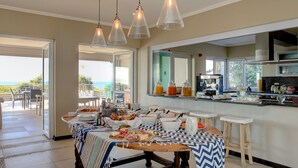 A breakfast setup with a buffet table, bar stools, and a kitchen area with a large window overlooking the outdoors. - Abalone Guest Lodge (Hermanus)