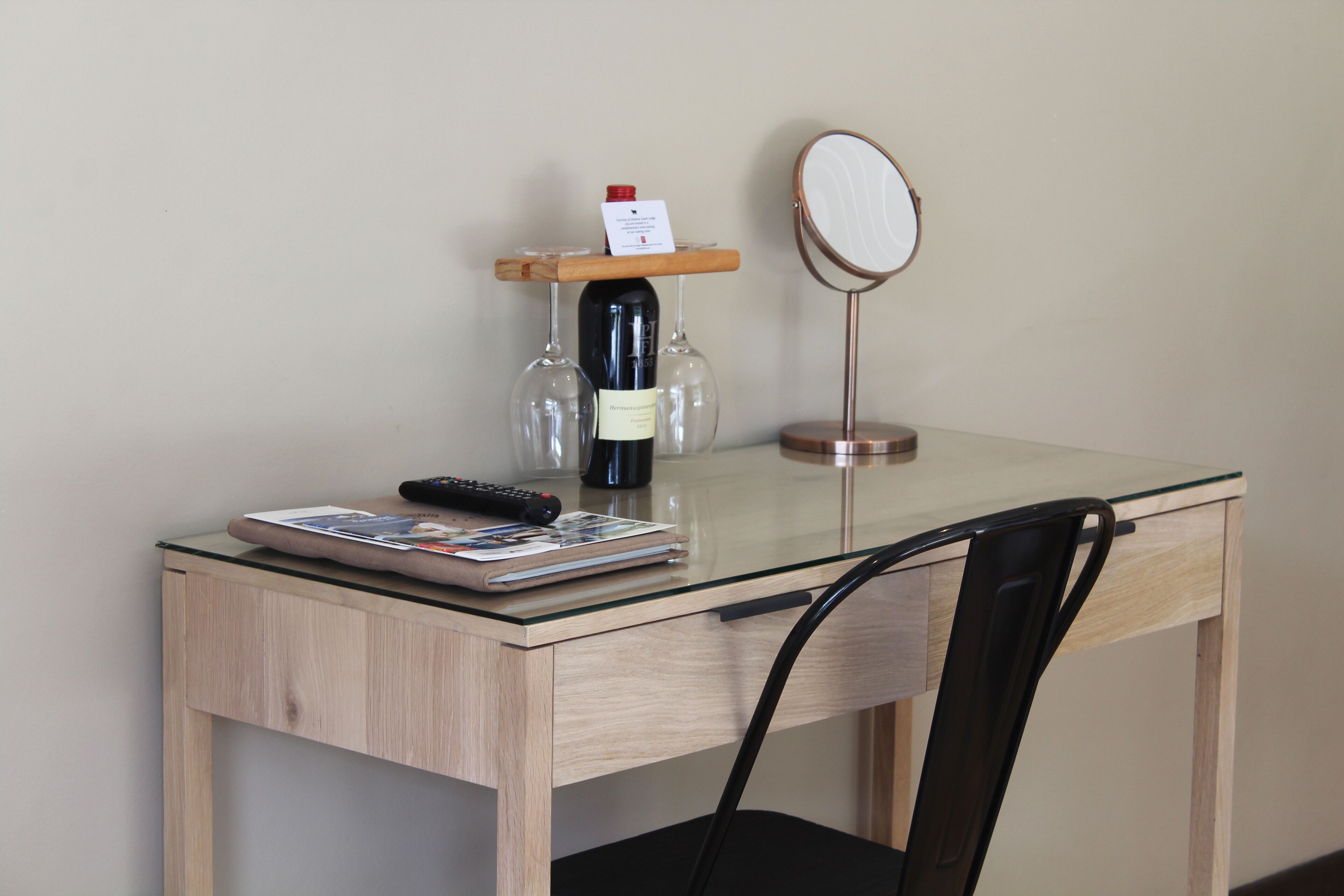 Una scrivania in legno con piano in vetro, una bottiglia di vino, due bicchieri da vino, un telecomando e una rivista.