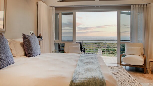 A spacious bedroom with a large bed, a view of the sea, and a balcony with a chair. - Abalone Guest Lodge (Hermanus)