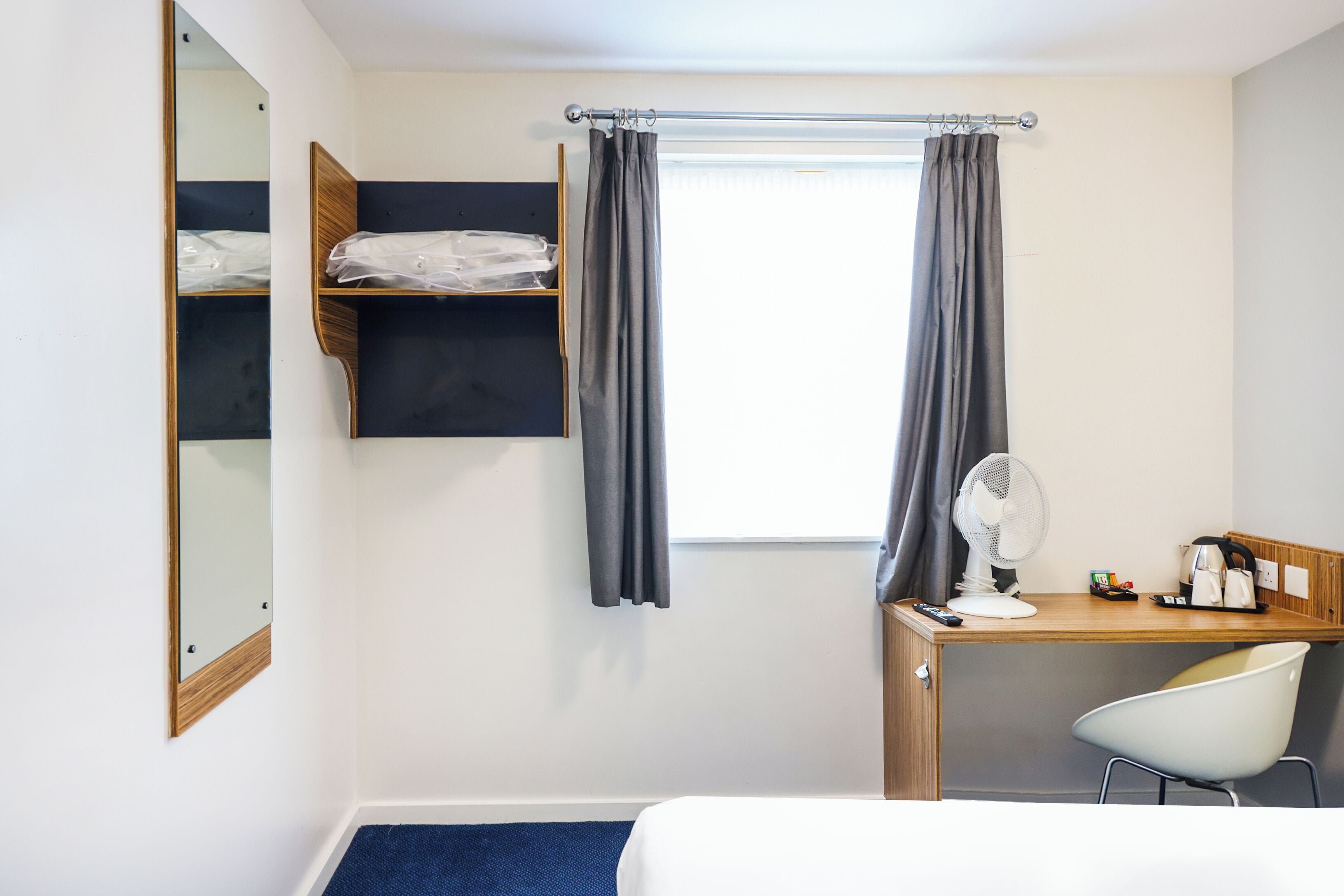 Une chambre d’hôtel avec un lit, un bureau et une chaise, un ventilateur et une fenêtre avec des rideaux.