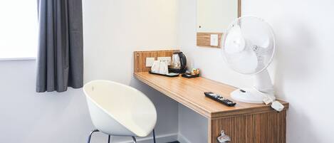 Habitación de hotel con escritorio de madera, una silla blanca, ventilador blanco, cafetera y espejo en la pared.