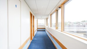 A corridor with a blue carpet, wooden doors, and large windows offering a view of buildings and a bridge.