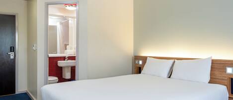 A hotel room with a large bed, a wooden headboard, a nightstand, a bathroom with a sink and toilet, and a door to the left.