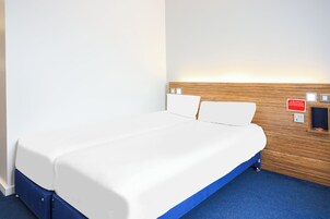 A hotel room with a bed, a wooden headboard, and a nightstand with a lamp.