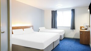 A hotel room with two beds, a wooden headboard, a blue carpet, a window with curtains, and a small wooden cabinet with a fan.