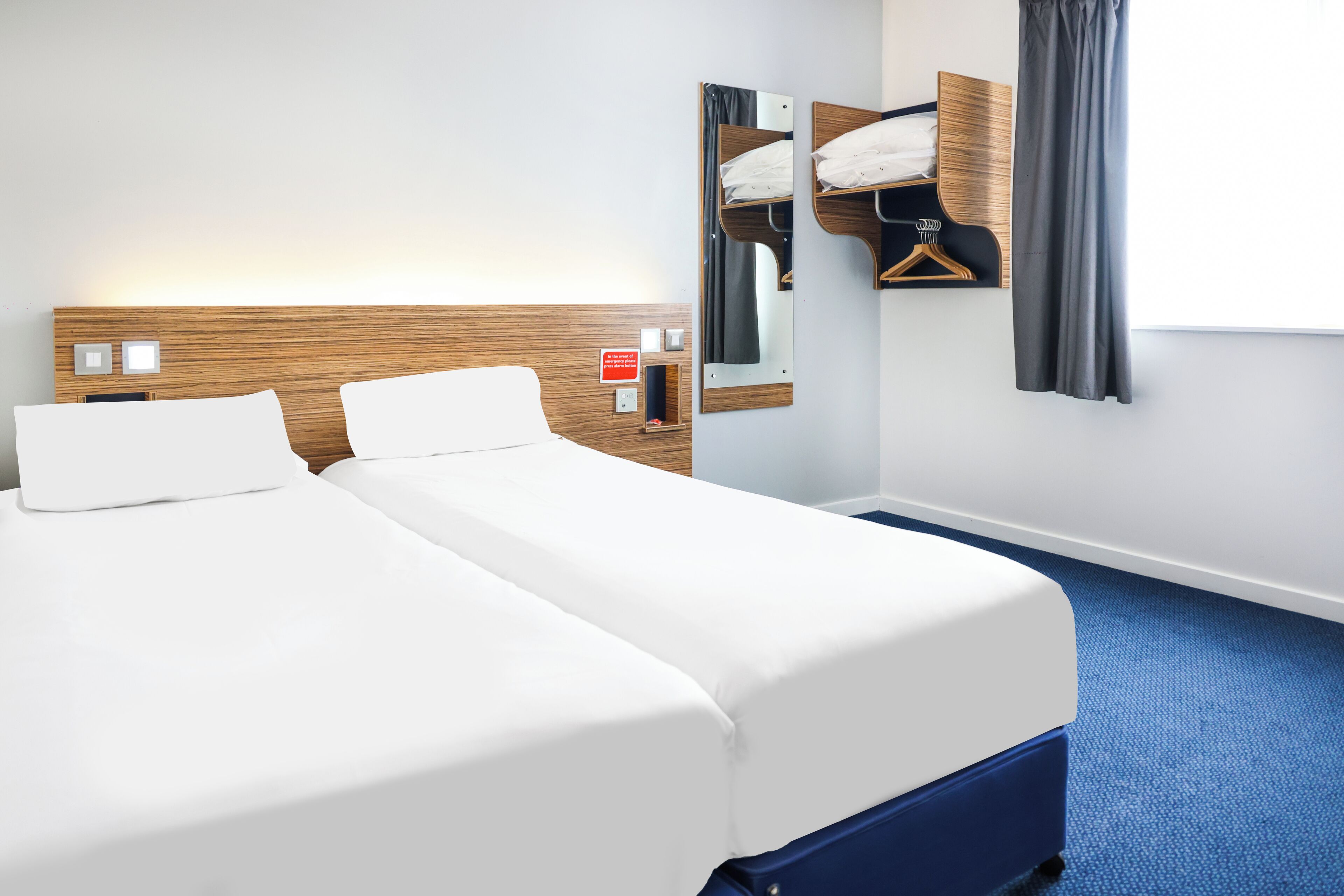 Ett hotellrum med två sängar, en sänggavel i trä, en spegel, en klädstång och en blå matta.