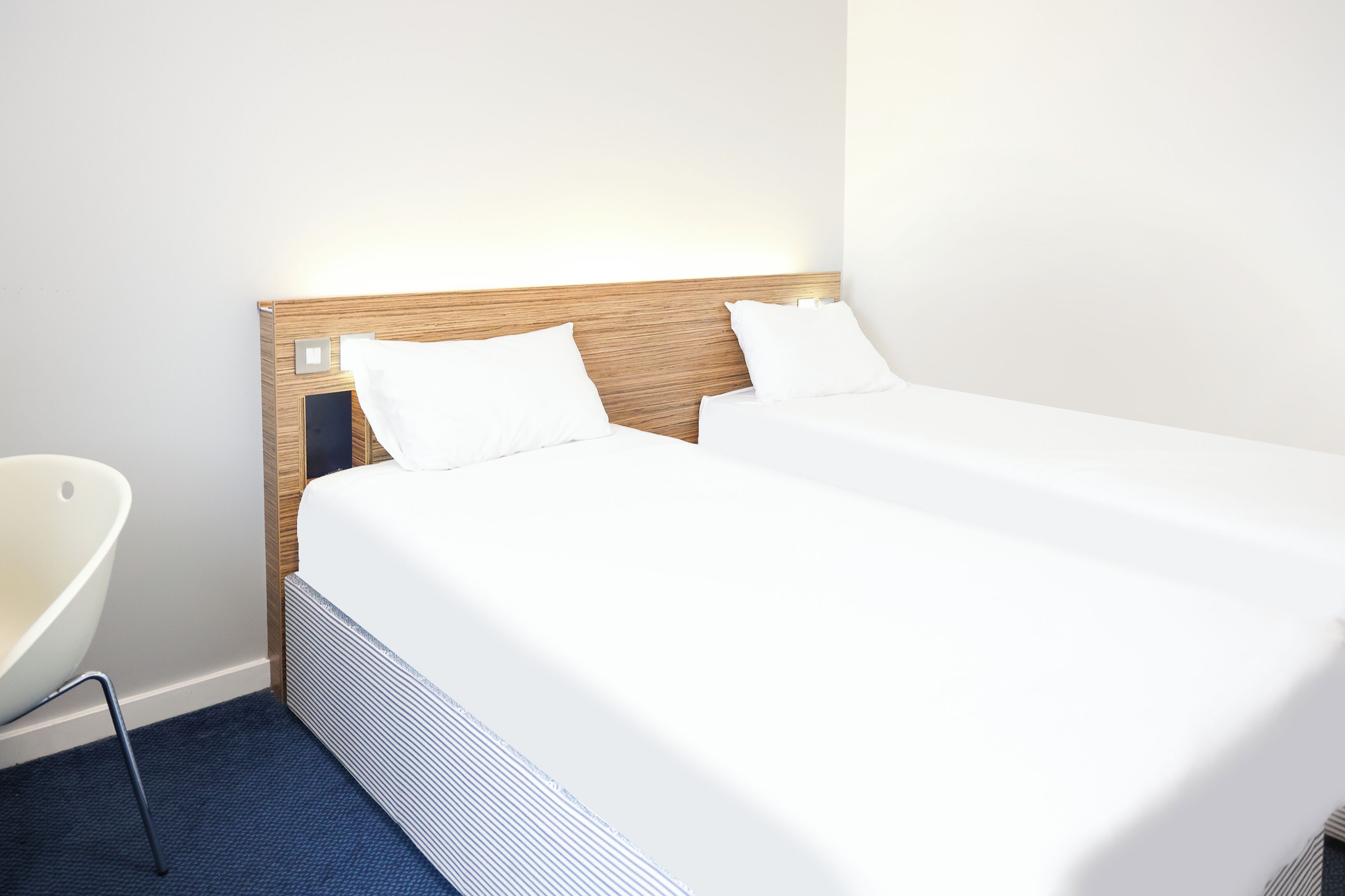 A neatly made twin bed with a wooden headboard and white bedding, a modern chair, and a dark blue carpet.