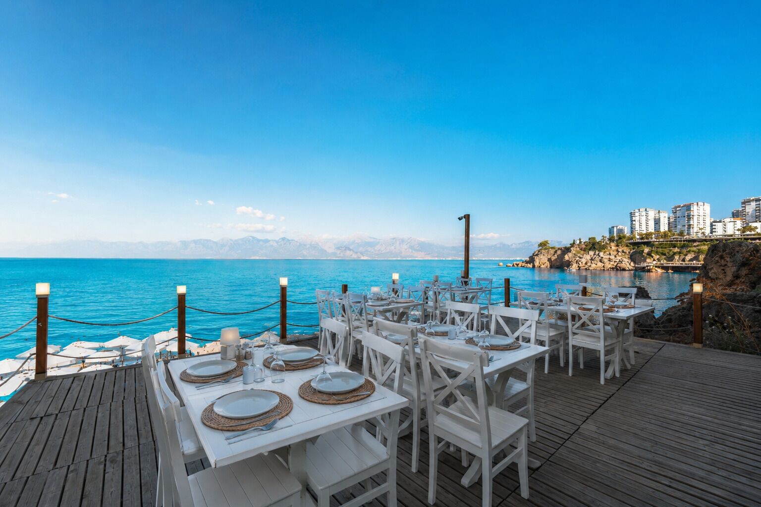 Un restaurante frente al mar, con mesas dispuestas para comer, con vistas a un mar azul y un paisaje montañoso.