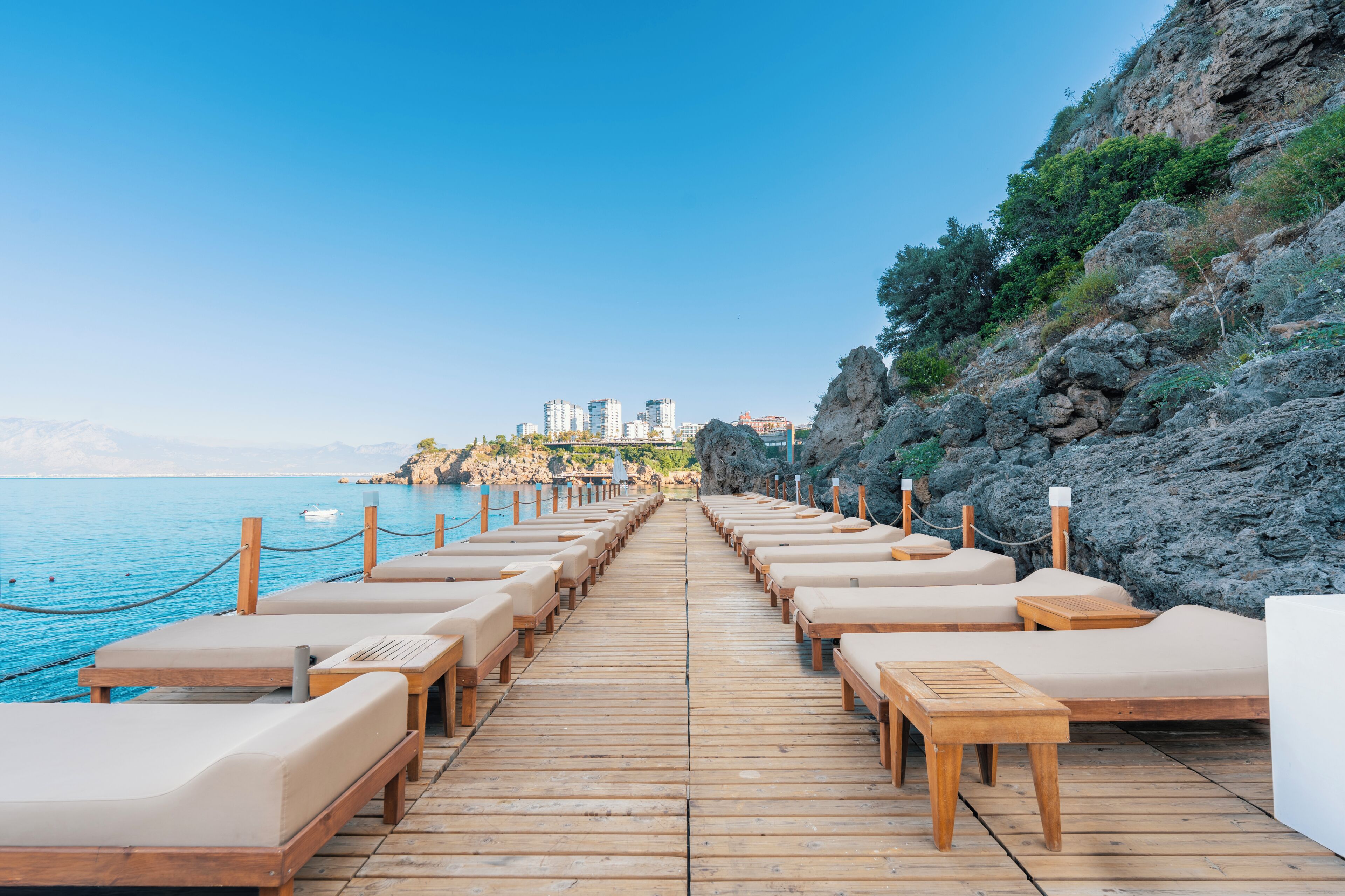 A wooden deck with lounge chairs and tables facing a coastal area with buildings in the background.