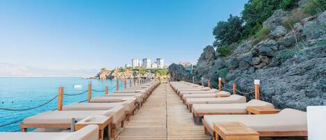 A wooden deck with lounge chairs and tables facing a coastal area with buildings in the background.