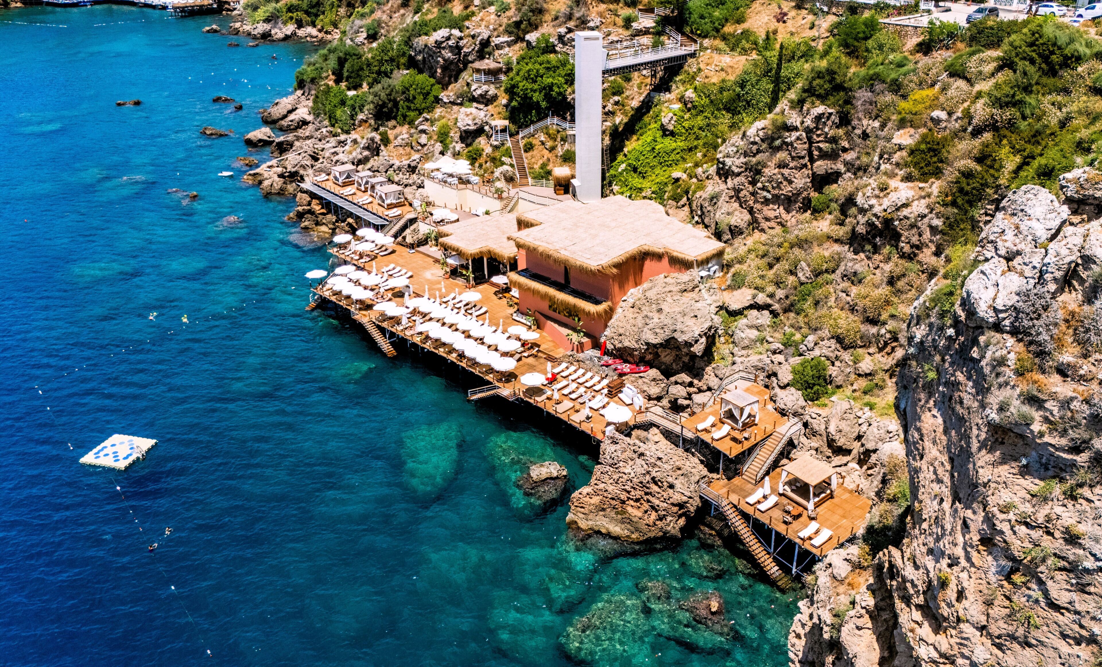 Eine Luftaufnahme eines Küstenhotels mit einer Pier, die ins Meer führt, mit Liegestühlen und einer Struktur mit Reetdach.