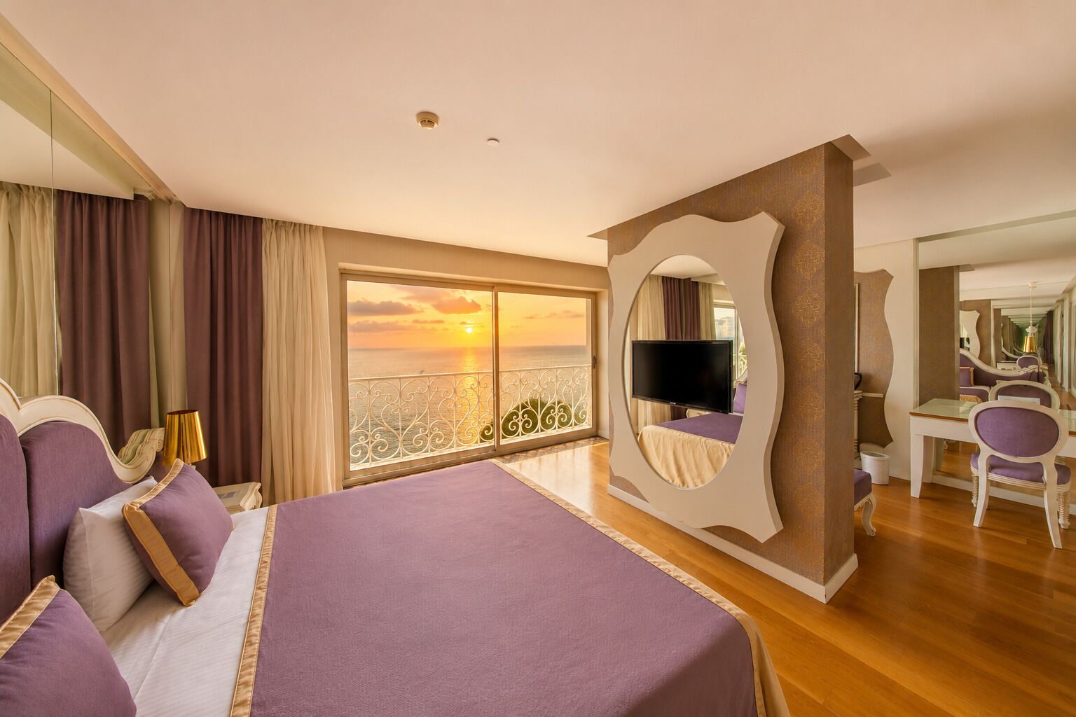 Habitación de hotel con una cama grande, televisión, escritorio con silla y balcón con vistas al atardecer.
