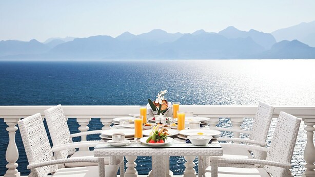 A dining table with white wicker chairs on a balcony overlooking a body of water with mountains in the distance.