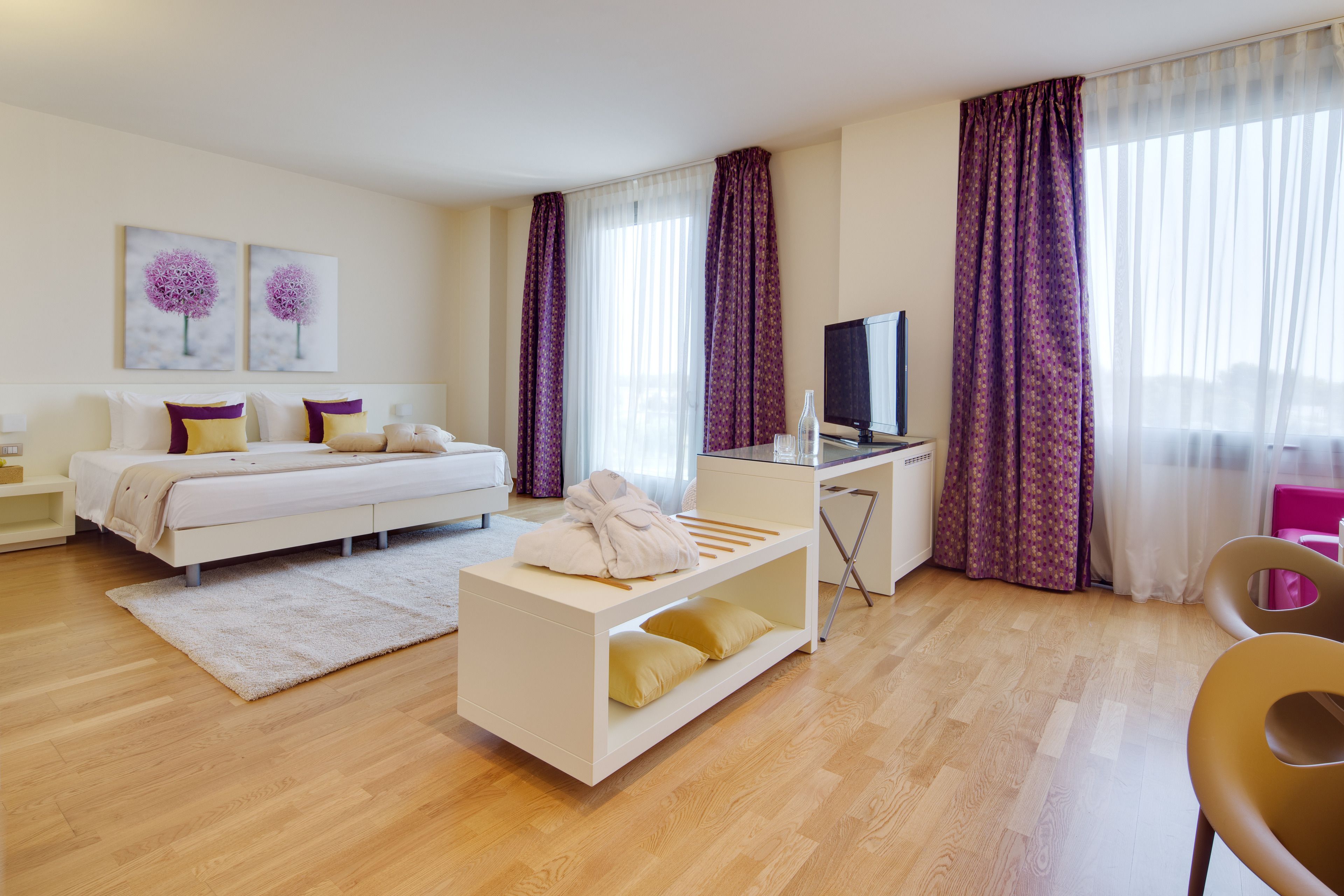 Una habitación de hotel moderna con una cama, un escritorio, un televisor y ventanas grandes con cortinas moradas.