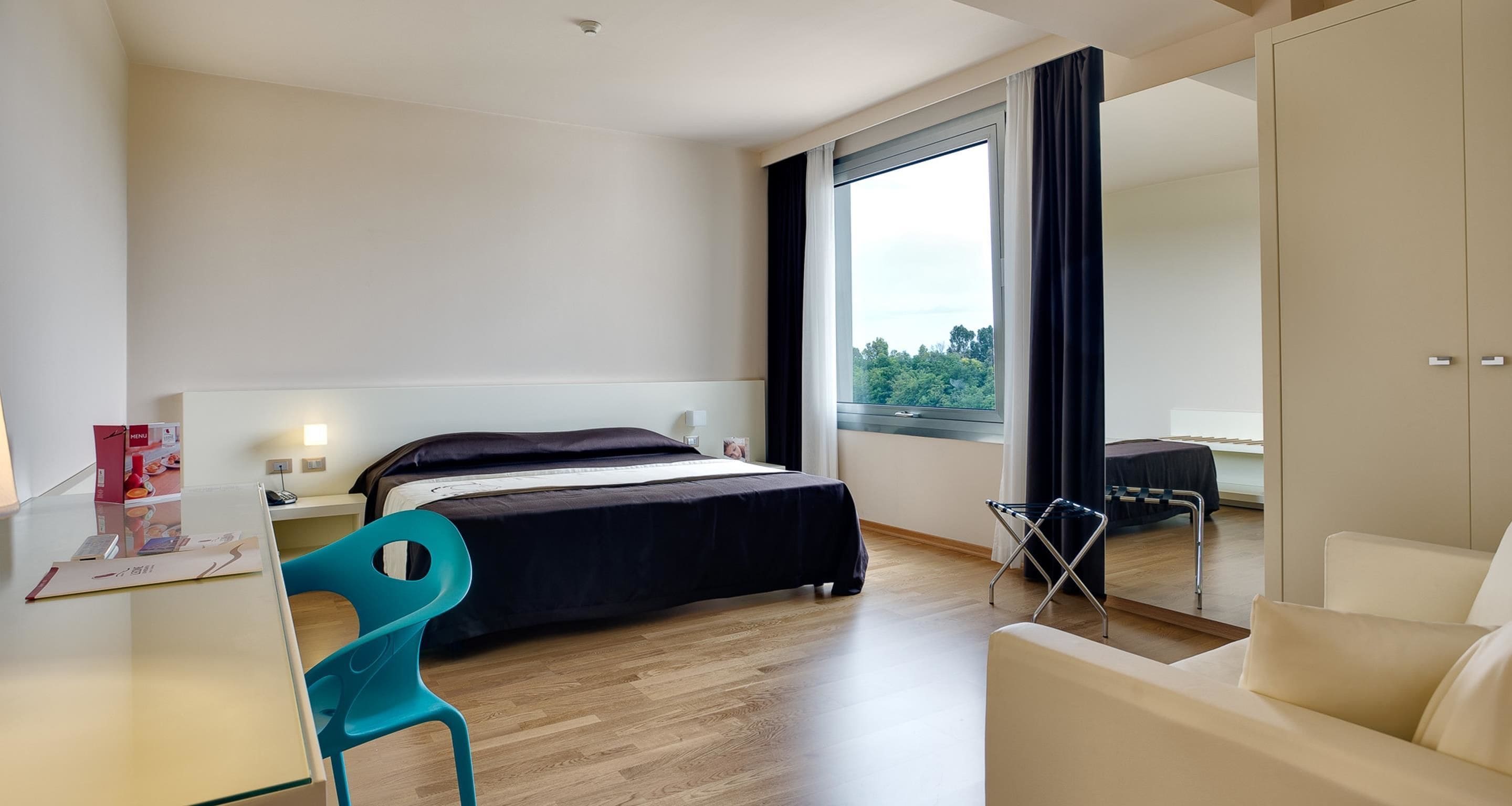 A modern hotel room with a bed, a desk, a chair, and a wardrobe.