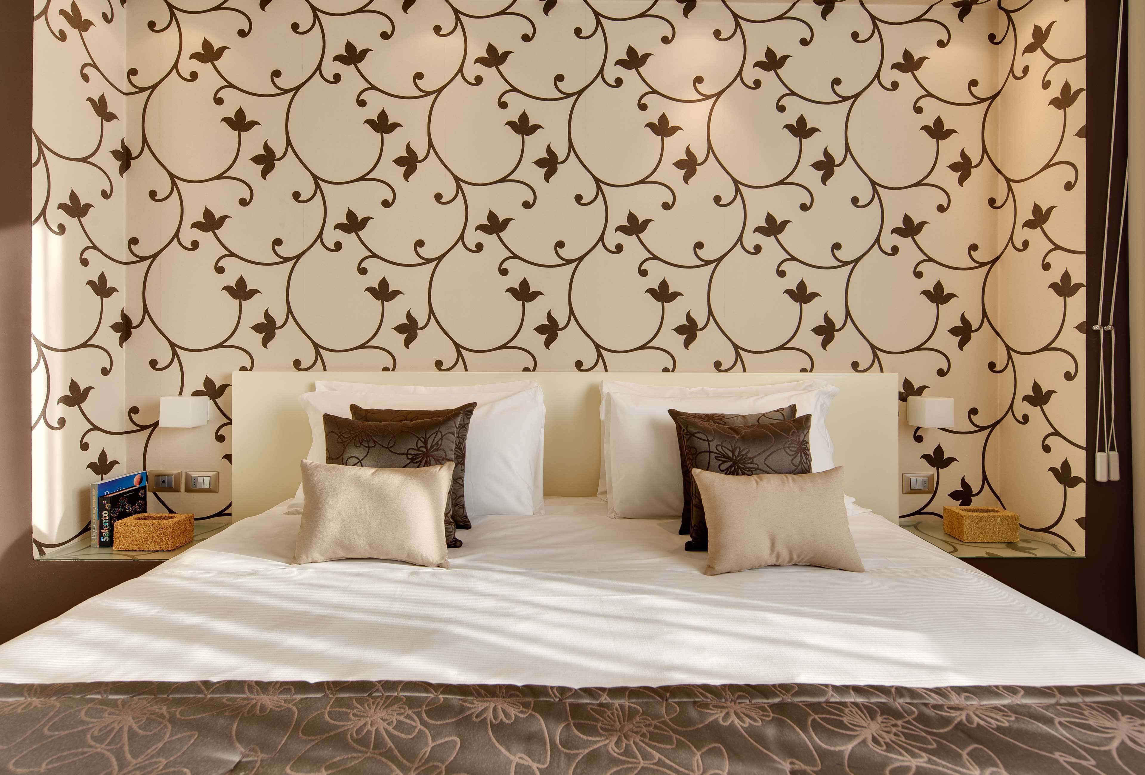Una cama bien hecha con sábanas blancas y almohadas con estampado floral marrón, una mesita de noche con lámpara y una pared con papel tapiz decorativo con motivos florales.