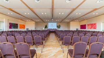 Een vergaderzaal met rijen paarse stoelen, een scherm aan de voorkant en houten balken in het plafond.