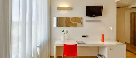 A modern hotel room with a white desk, a red chair, a TV mounted on the wall, and a vase with flowers on the desk.