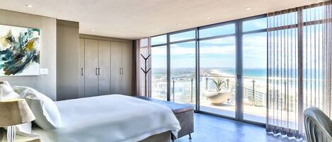 A modern bedroom with a large bed, a view of the beach, and a cityscape through floor-to-ceiling windows.