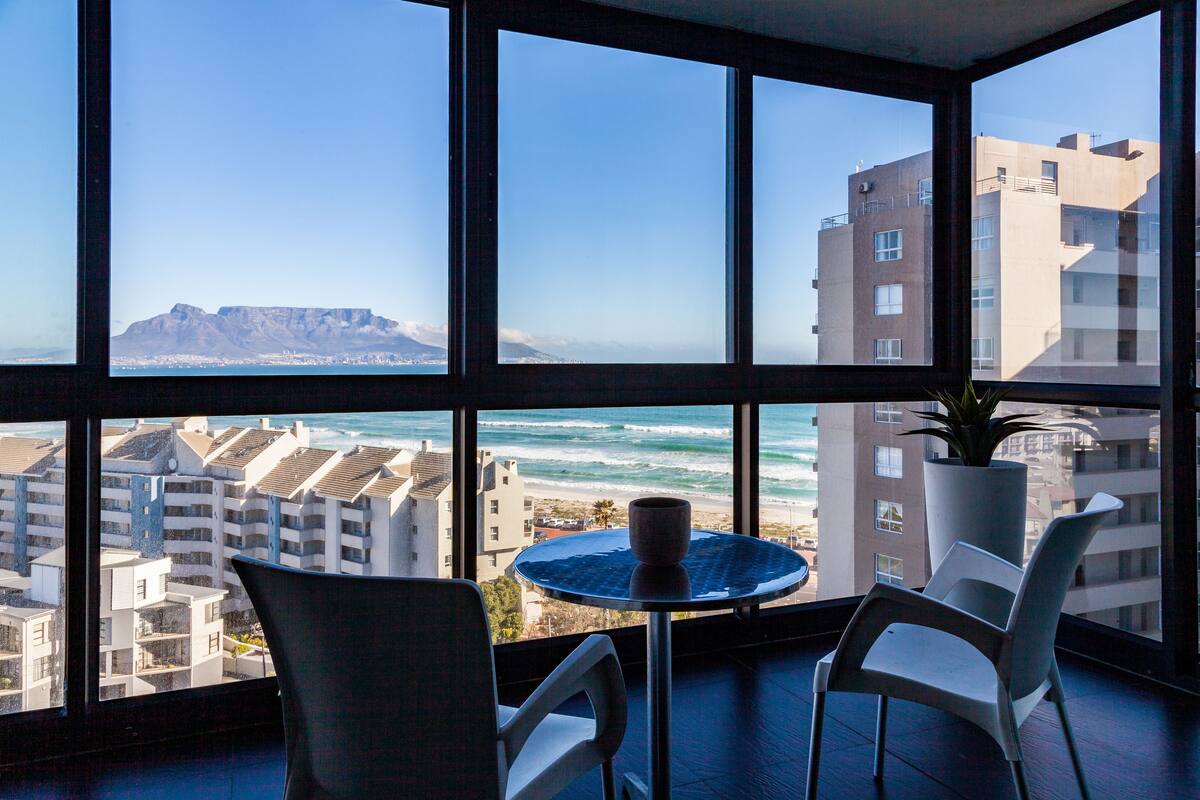 Quarto com uma grande janela com vista para o oceano, uma mesa redonda com duas cadeiras e um vaso de planta.