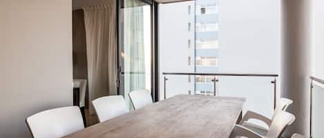 A modern dining area with a wooden table and white chairs, a glass partition, and a view of a building through the window.