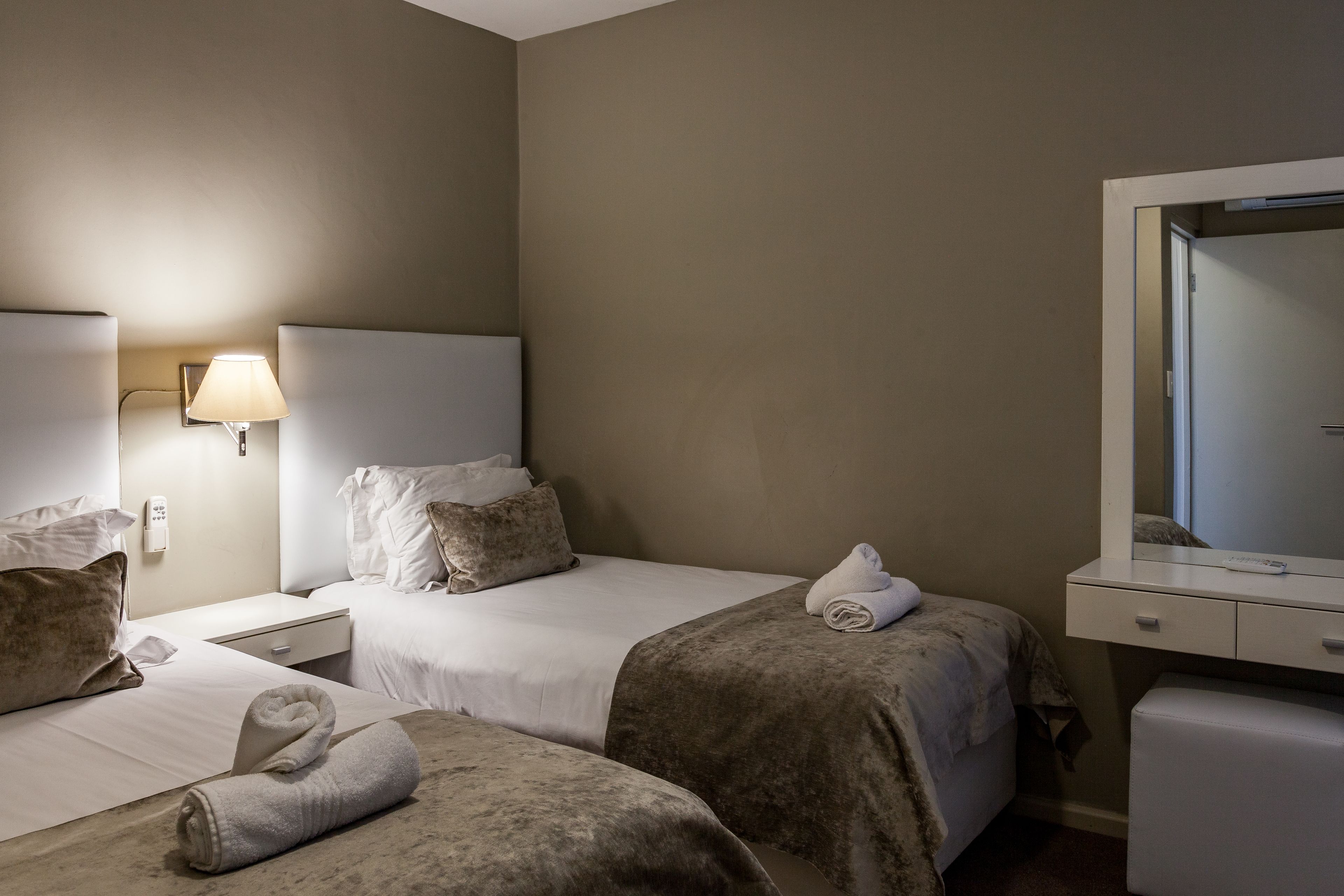 A hotel room with two beds, a bedside table with a lamp, a dresser with a mirror, and a small seating area.