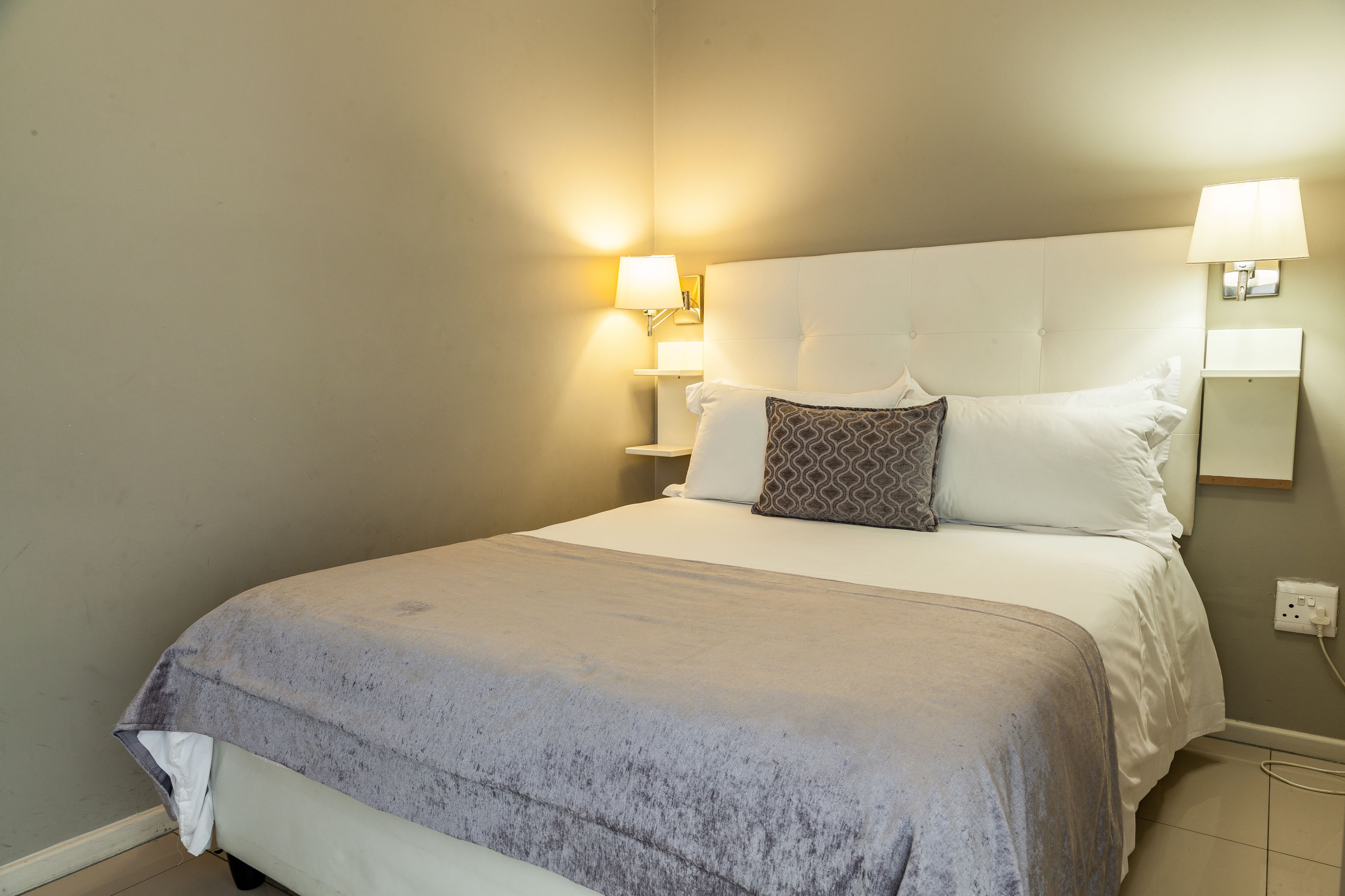 Ein ordentlich bezogenes Bett mit grauer Decke, zwei Nachttischlampen und einer Wandleuchte.