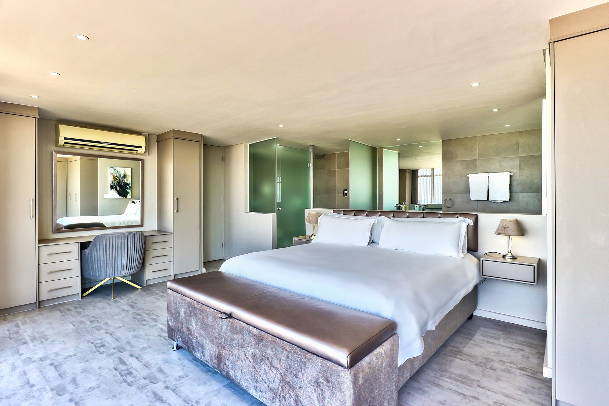 A modern hotel bedroom with a large bed, a bench, a vanity with a mirror, and a walk-in closet.