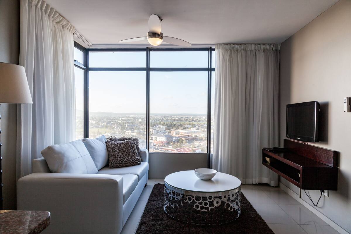 Sala de estar moderna com sofá, mesa de centro redonda, televisão e uma grande janela com vista para a cidade.