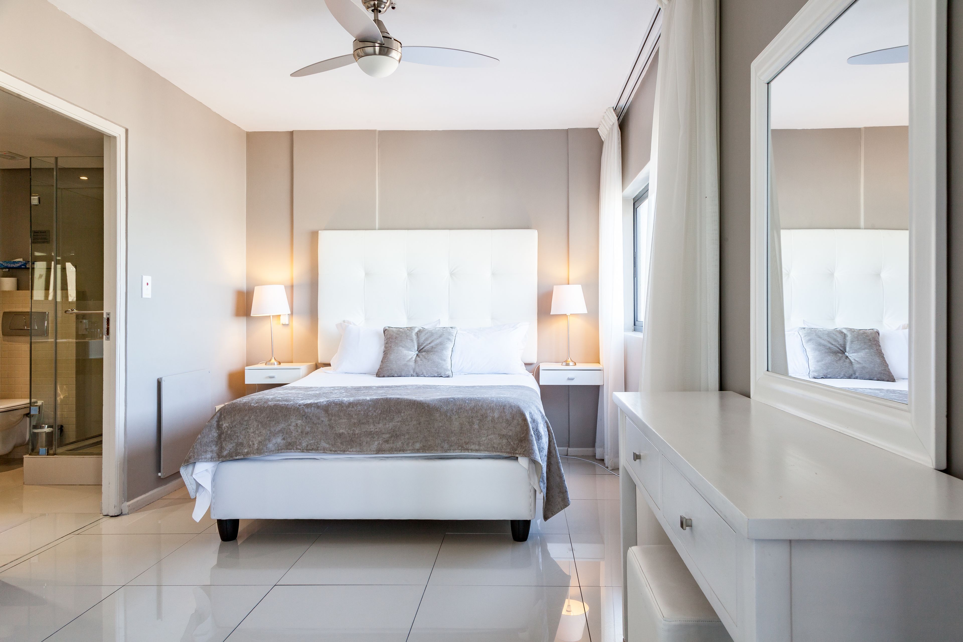 A modern hotel bedroom with a large bed, bedside lamps, a ceiling fan, and a large mirror.