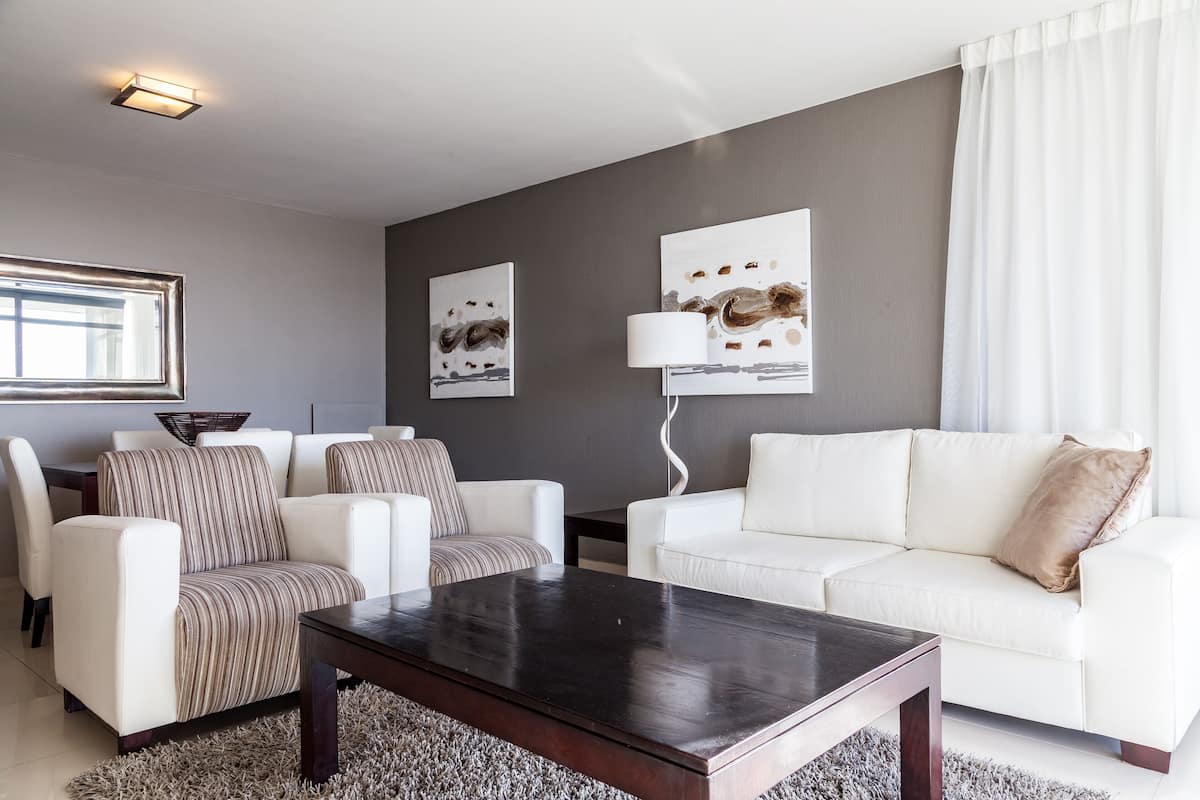 Uma sala de estar moderna com uma mesa de centro de madeira escura, sofás brancos e almofadas listradas. Há duas pinturas abstratas na parede cinzenta e um abajur.