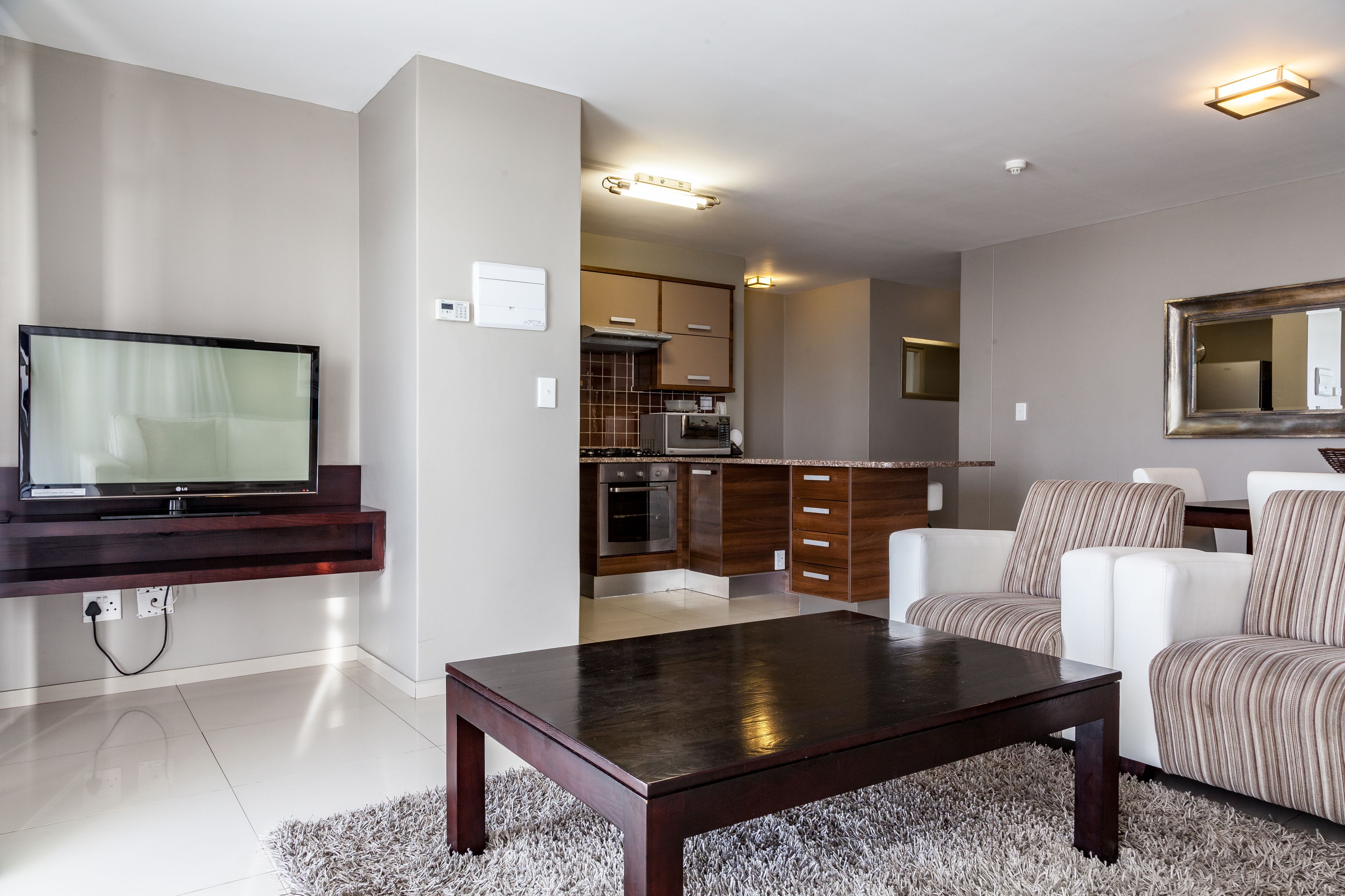 A modern living room with a flat-screen TV, a dark wooden coffee table, and a striped sofa. A kitchen is visible in the background with wooden cabinets and a microwave.