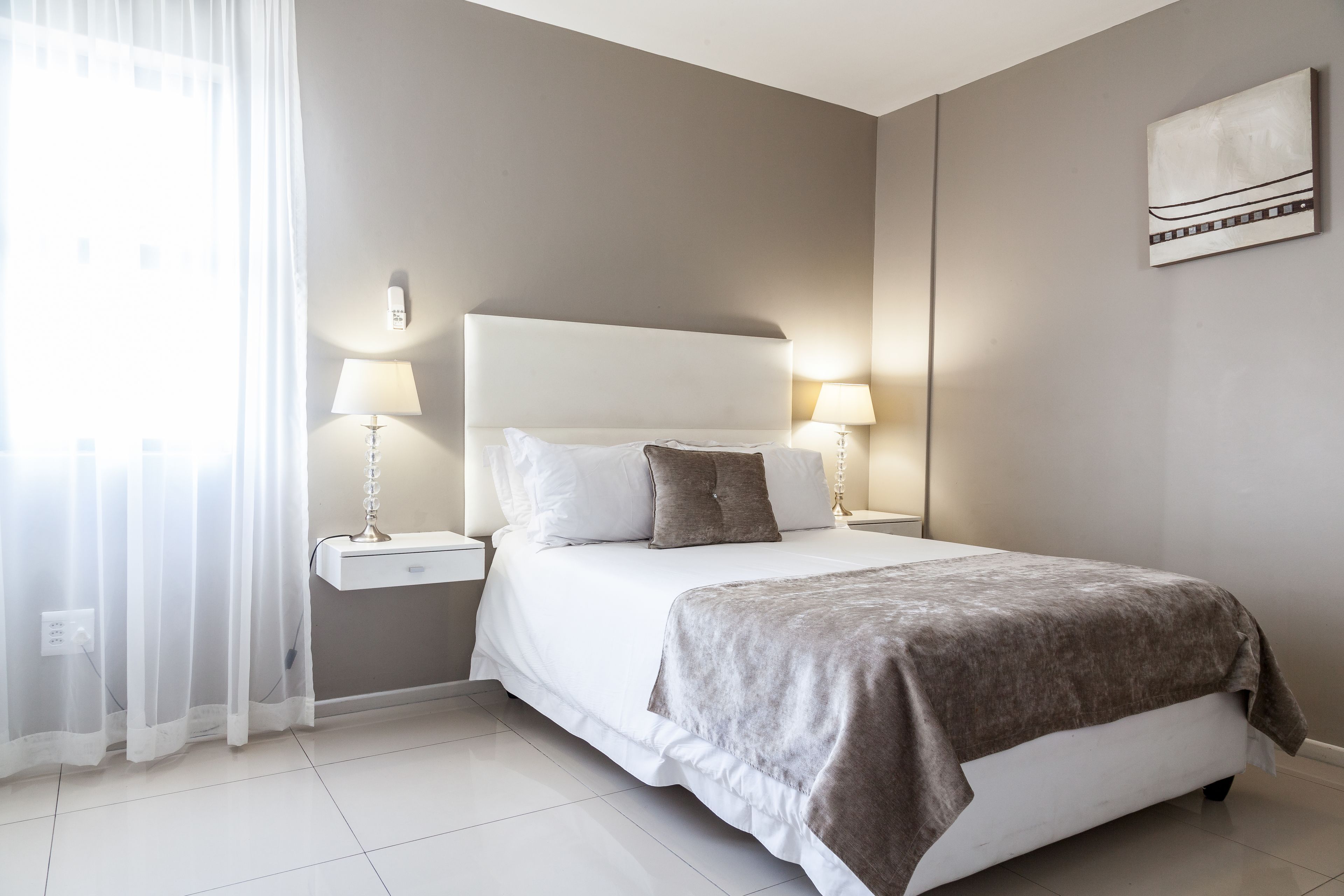 A neatly arranged bedroom with a large bed, two bedside tables with lamps, a window with sheer curtains, and a wall-mounted artwork.