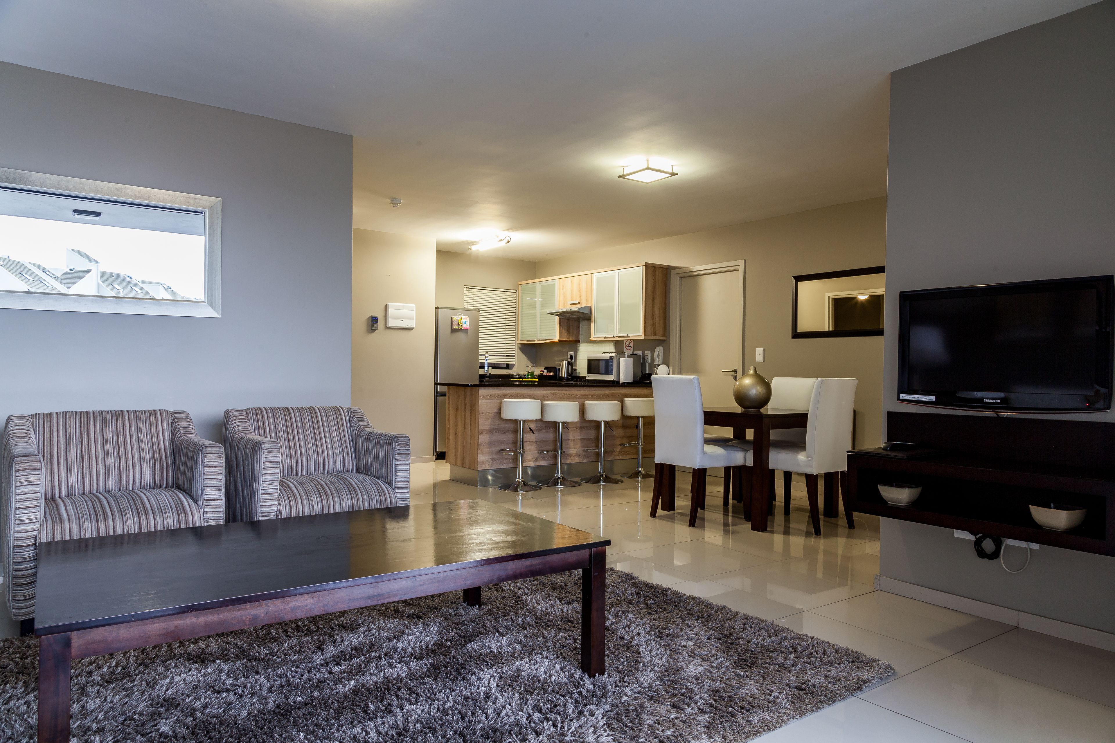 A living room with a sofa, coffee table, and TV. There is a kitchen in the background with a dining area.
