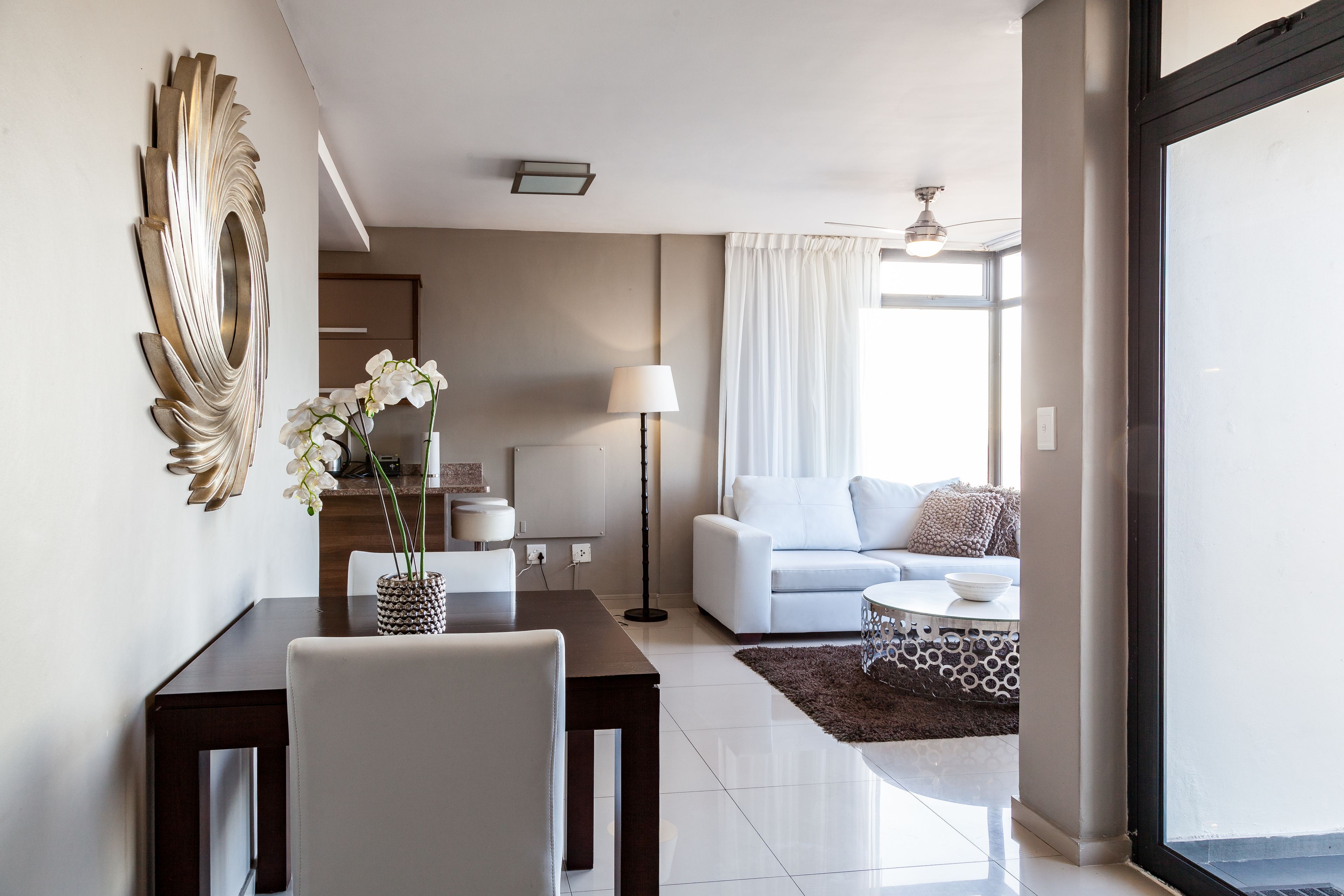 A modern living room with a white sofa, a dining table with chairs, a decorative mirror, and a vase with white flowers.