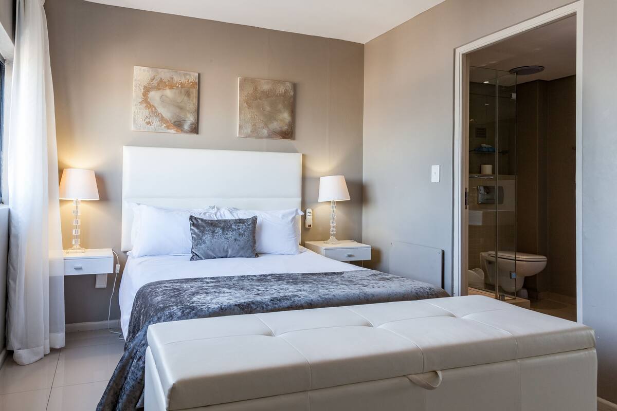Um quarto moderno com uma cama grande, duas mesas de cabeceira e um chuveiro com paredes de vidro.