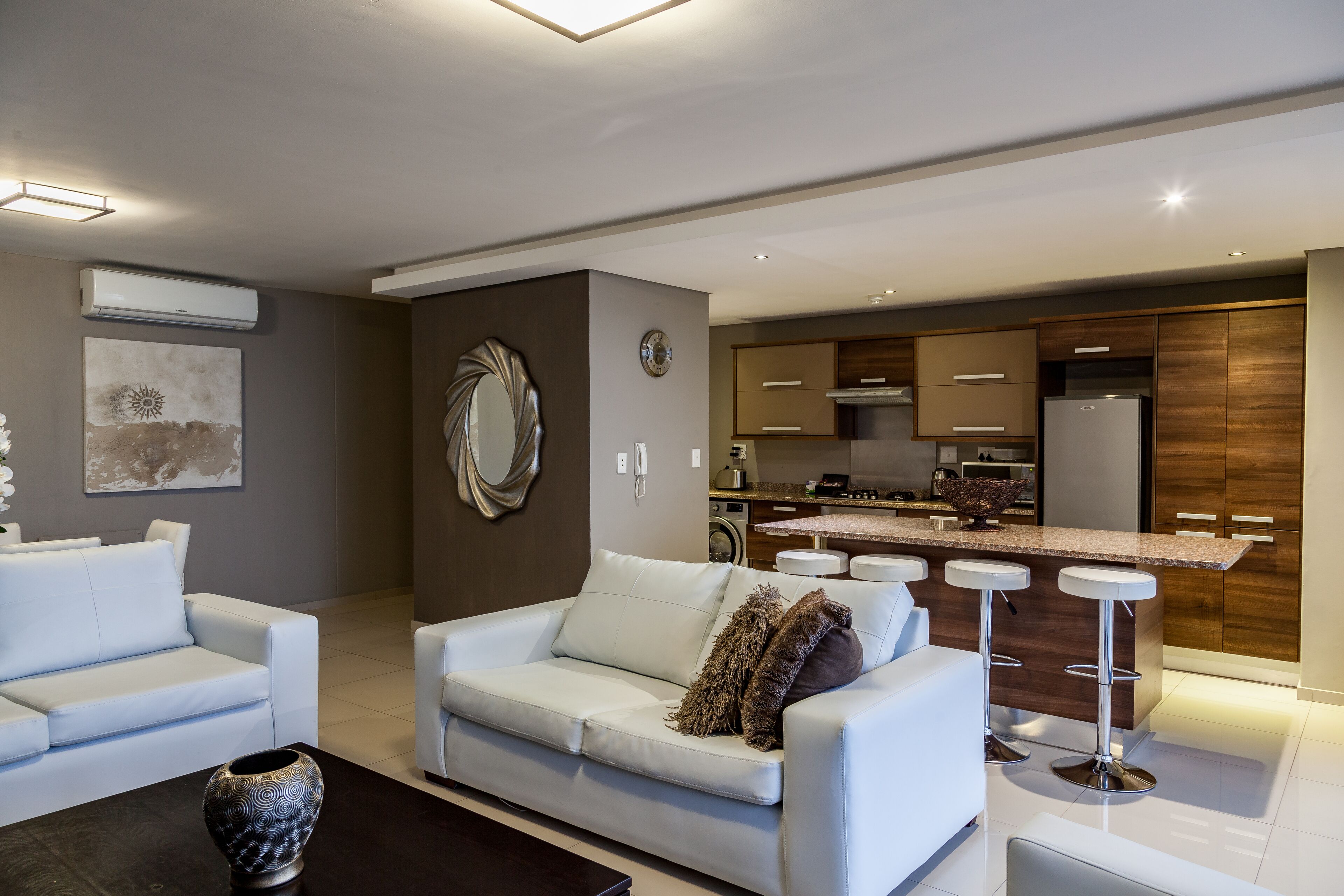 A modern living room with a white sofa, a coffee table, and a kitchen area with wooden cabinets and a stainless steel refrigerator.