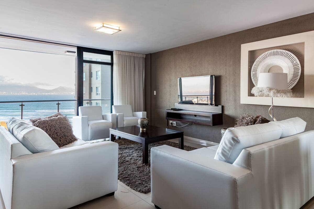 Uma sala de estar moderna com uma grande janela com vista para o mar, uma televisão de tela plana e um objeto decorativo na parede.