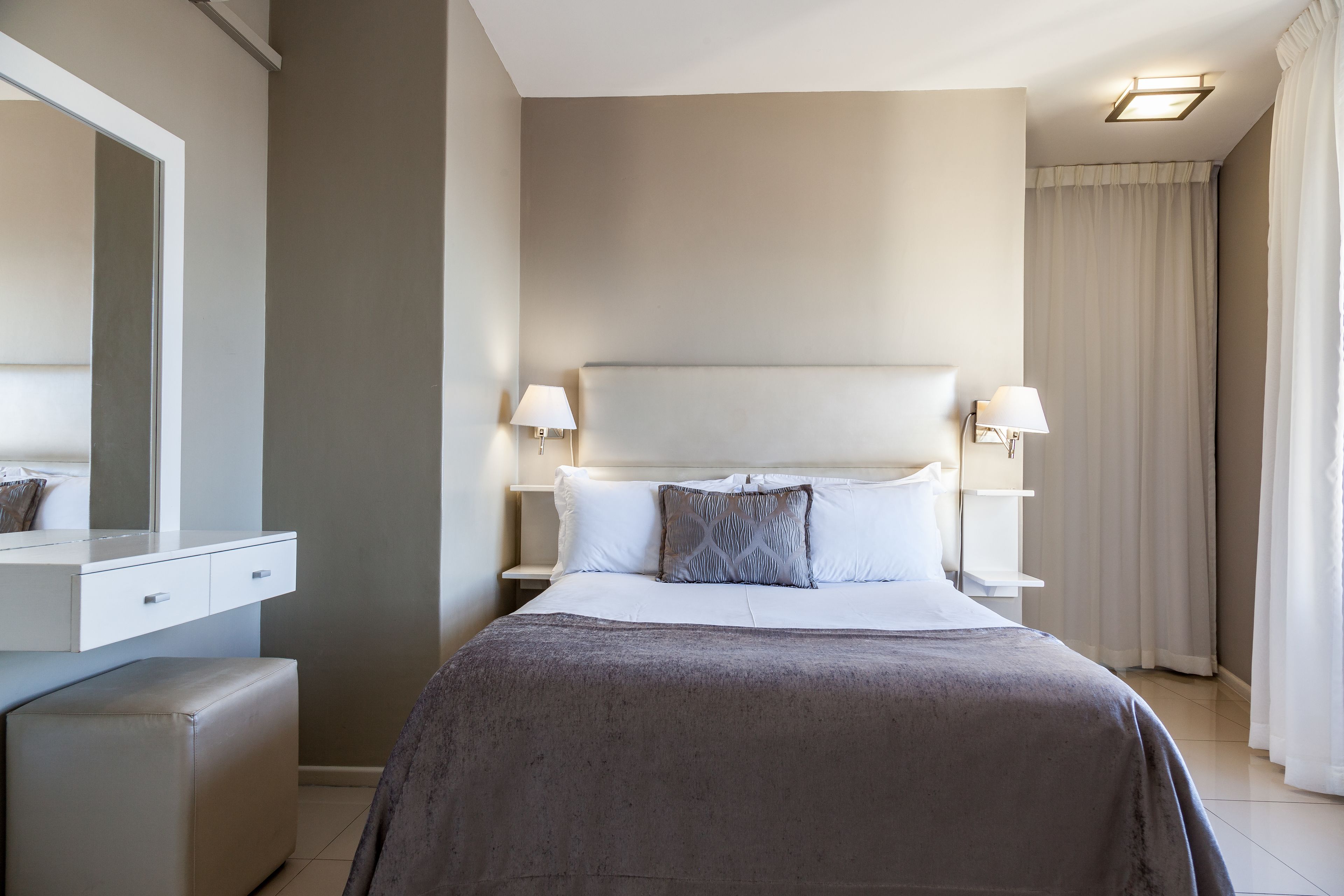Habitación de hotel con una cama grande, dos lámparas de noche, una mesita de noche, un espejo y una ventana con cortinas.