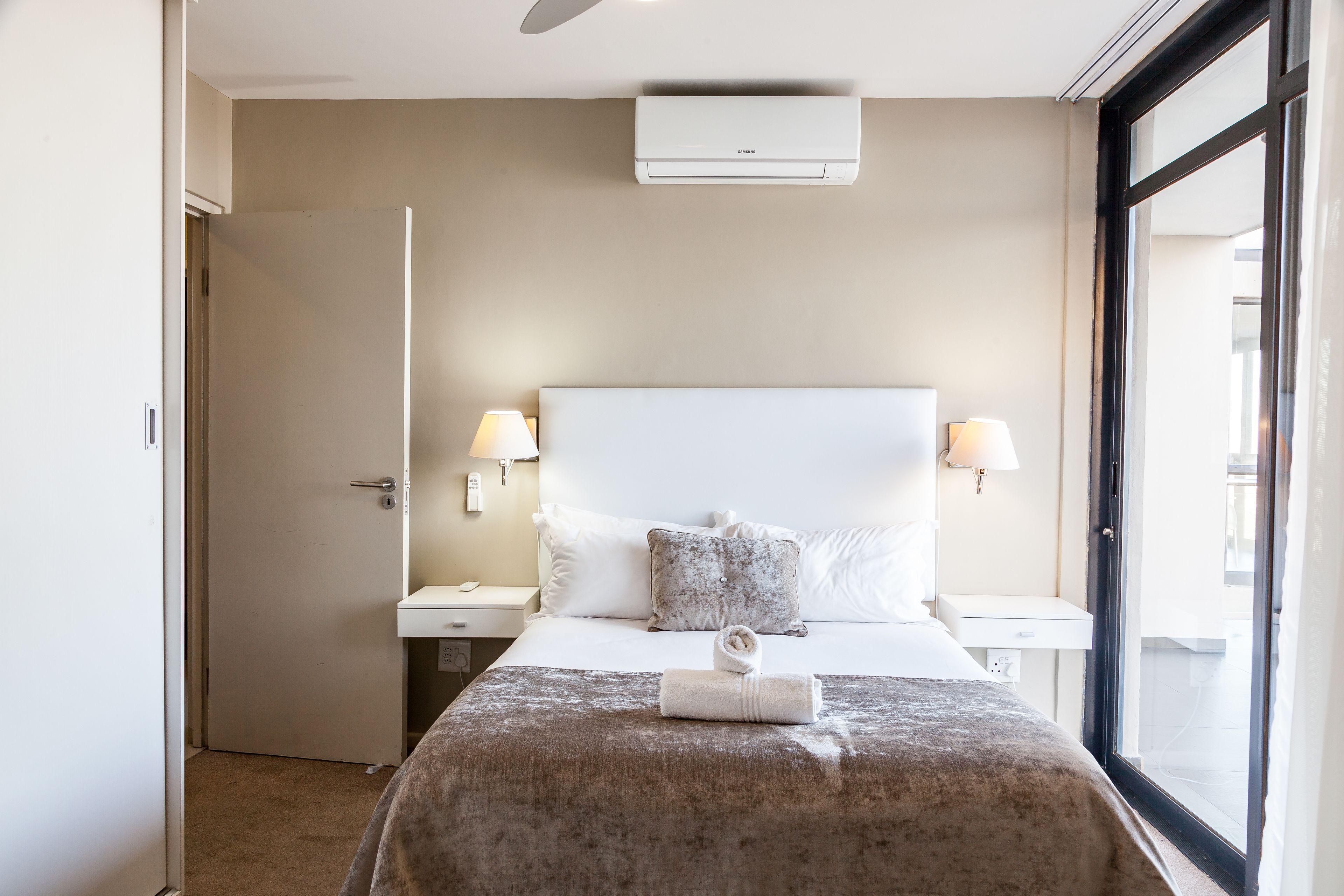 Una habitación de hotel con cama, lámparas de noche, una ventana y un equipo de aire acondicionado.