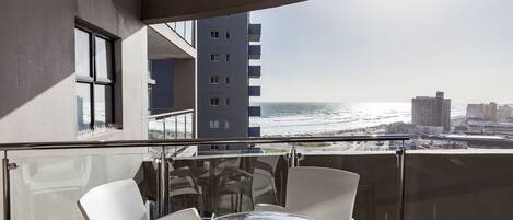 A balcony with a glass railing, white chairs, and a small round table. A tall building is visible in the background, and the ocean is in the distance.
