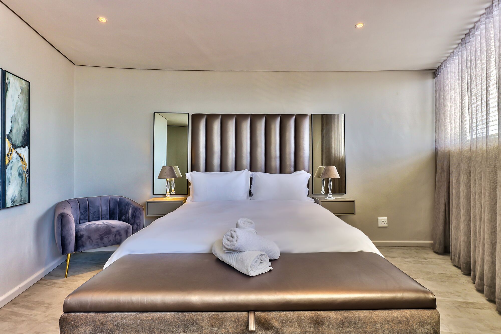 Een moderne hotelkamer met een groot bed, een nachtkastje met lampen, een stoel en een grote spiegel.