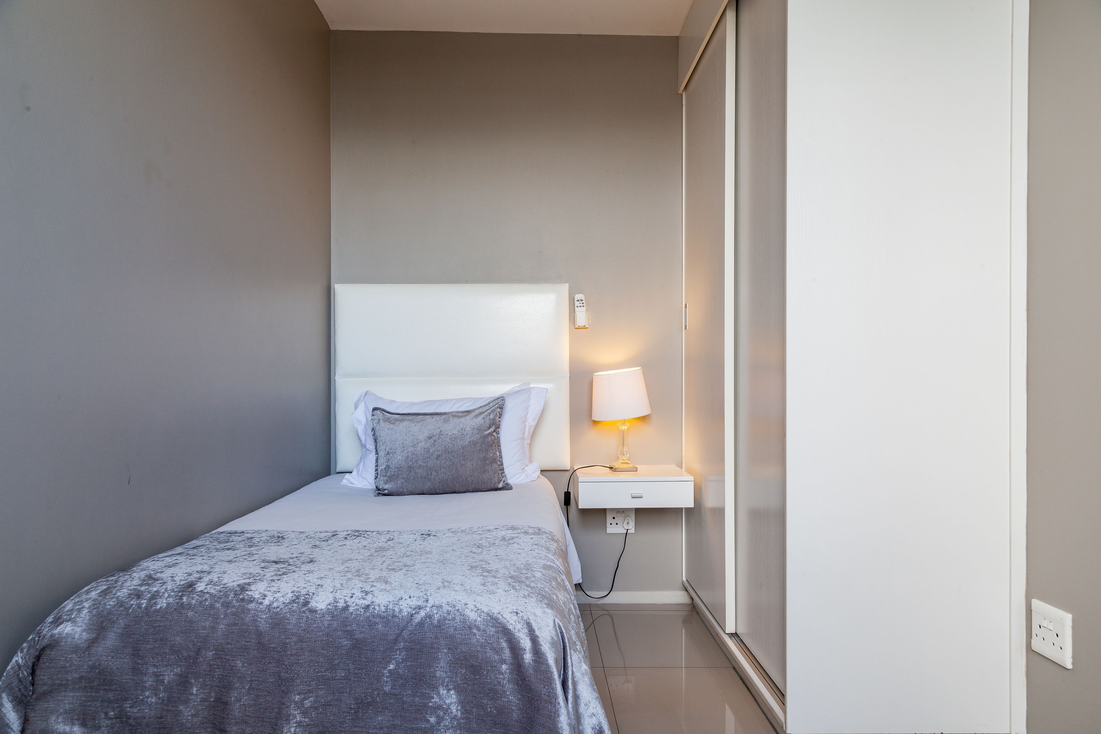 A small, neatly made bed with a grey blanket and pillows, a bedside table with a lamp, and a wall socket.
