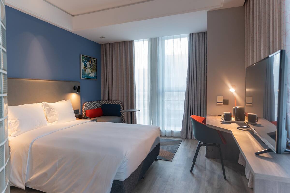 A hotel room with a large bed, a desk, a chair, a TV, and a window with curtains.