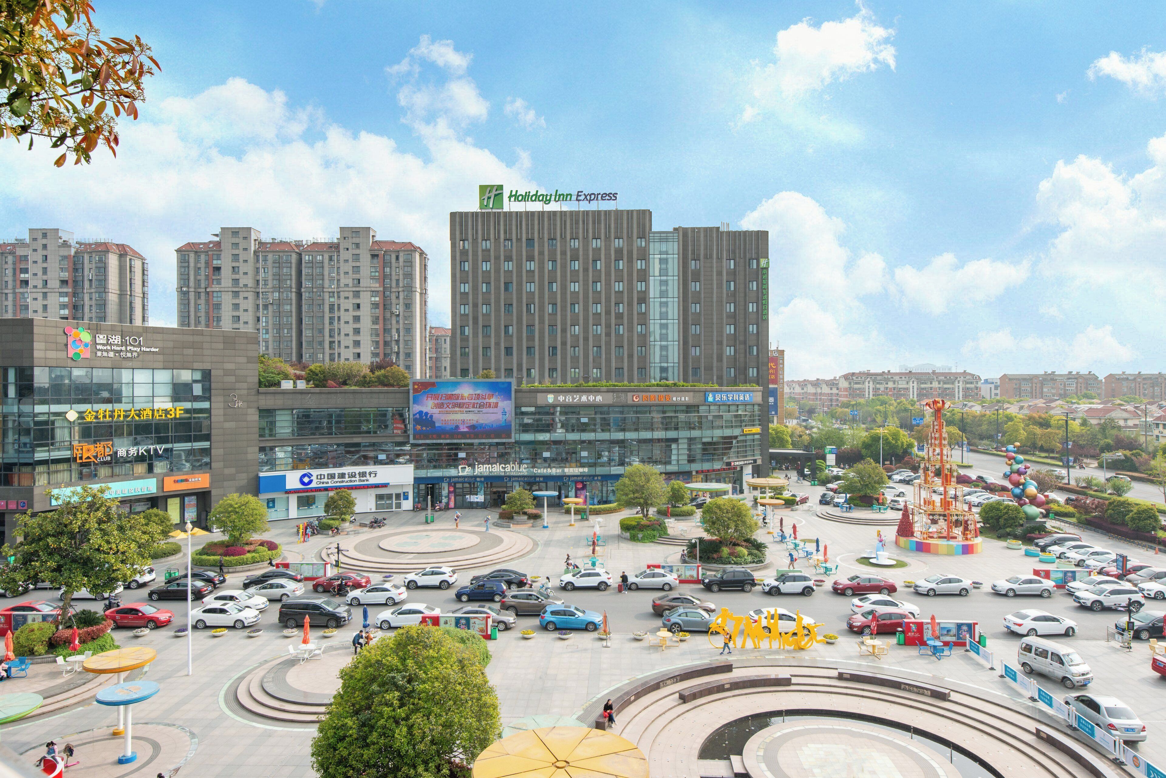 Un hotel de varios pisos con un llamativo letrero de Holiday Inn Express, rodeado por un estacionamiento con diversos vehículos y una plaza central con asientos y una escultura decorativa.