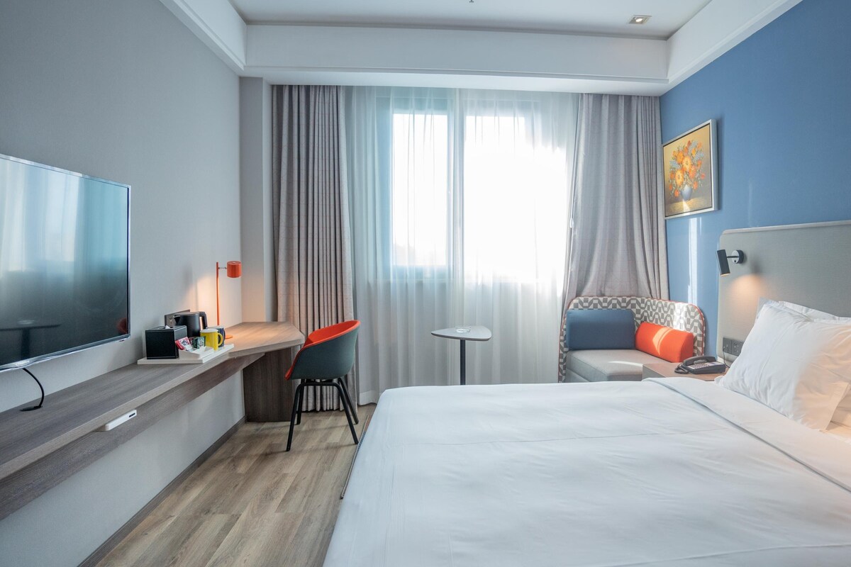 Une chambre d’hôtel avec un lit, un bureau et une chaise, une télévision, une petite table et un tableau au mur.